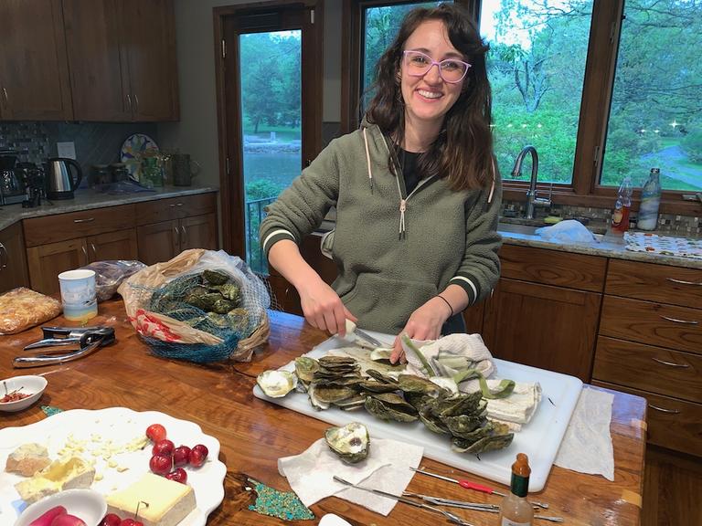 Shucking a tray of local oysters at the kitchen island, shucking knife in hand, with the mesh bag they came in alongside and the water visible through the windows behind.