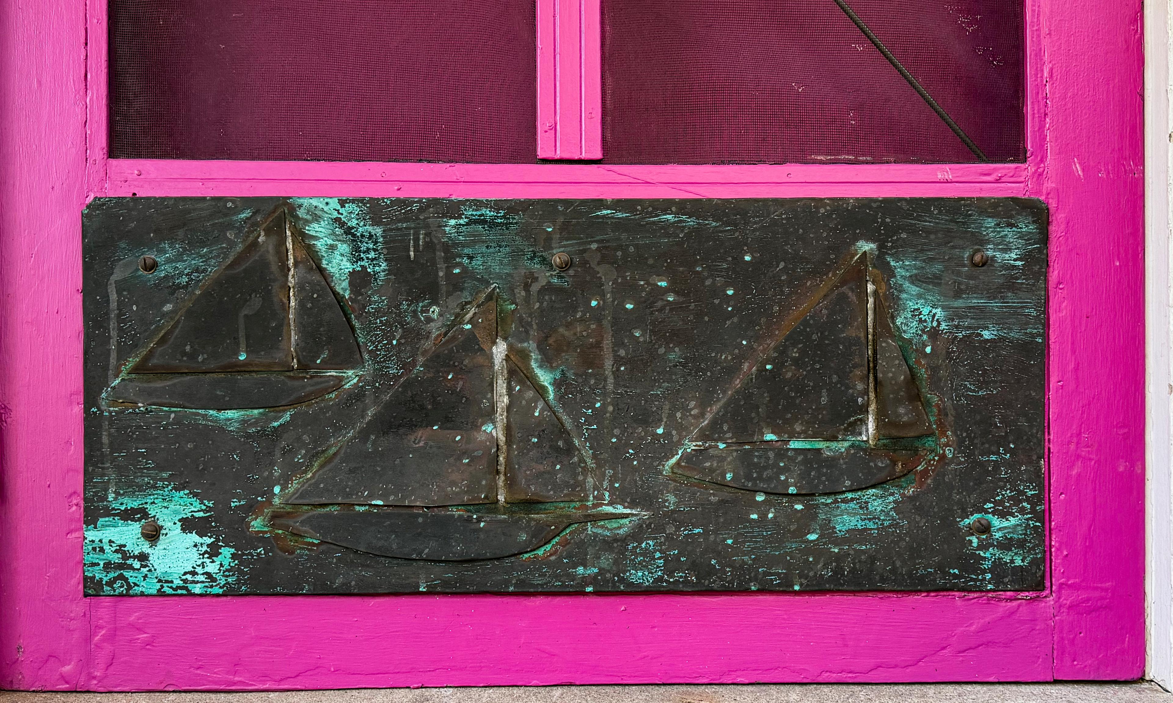 The sailboat kick plate mounted at the base of the blue screen door, weathered to turquoise verdigris