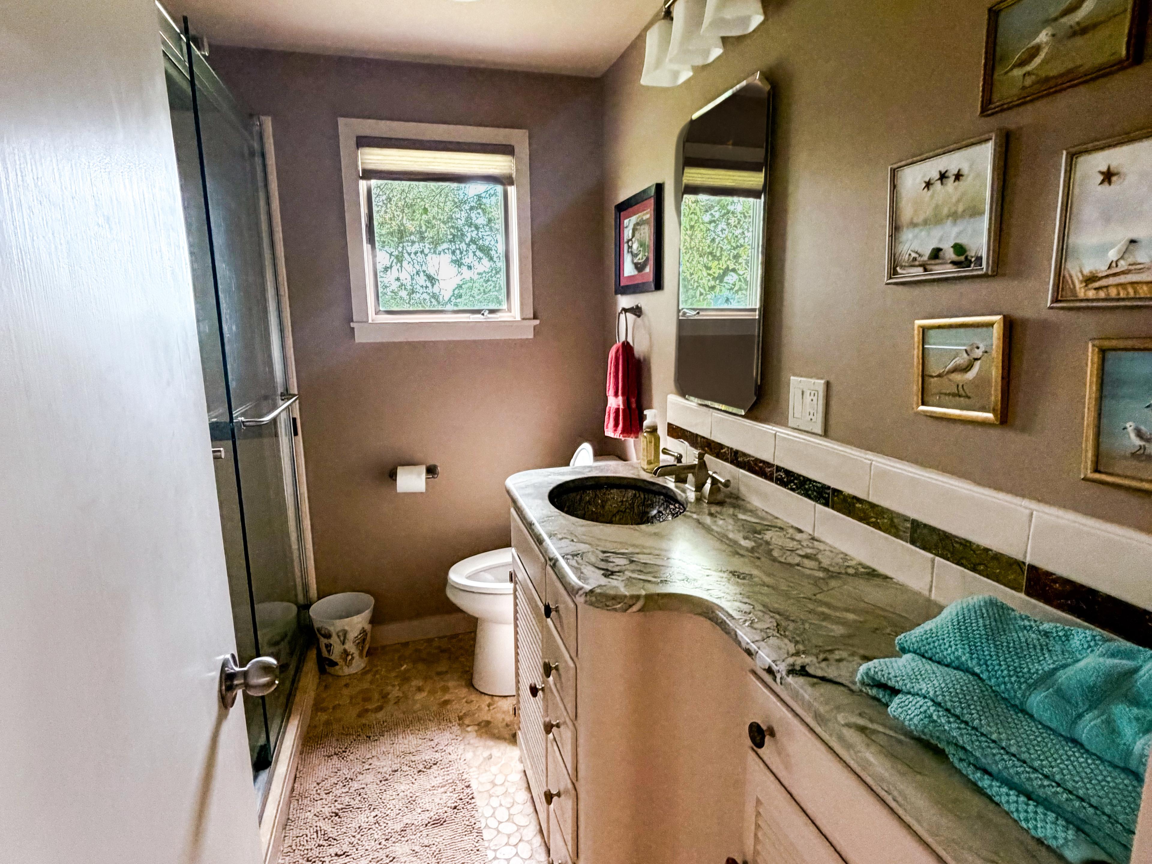 The master bath: pebble-stone floor, glass-door shower, and the curved granite vanity under a wall of shorebird paintings.