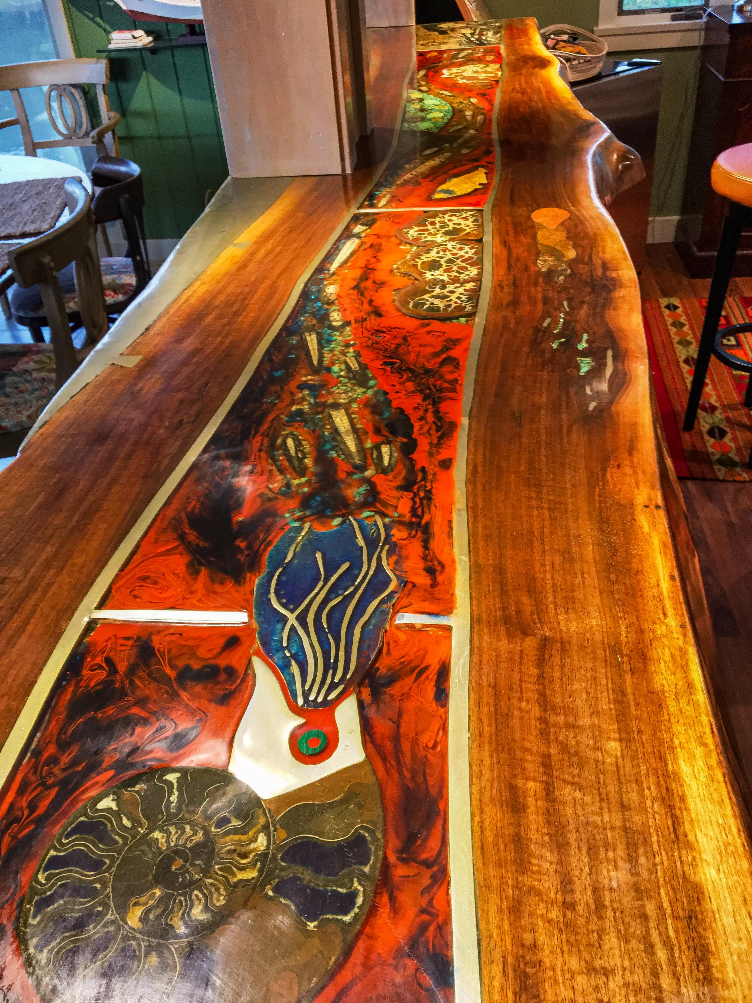 Looking down the length of the bar top: a river of orange resin set with an ammonite fossil, blue enamel, and turquoise, running between live-edge walnut slabs.