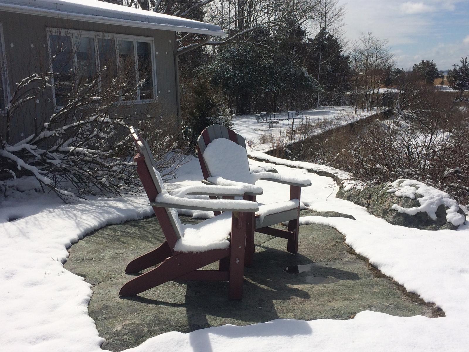 Two Adirondack chairs on the stone patio, each wearing a cushion of snow